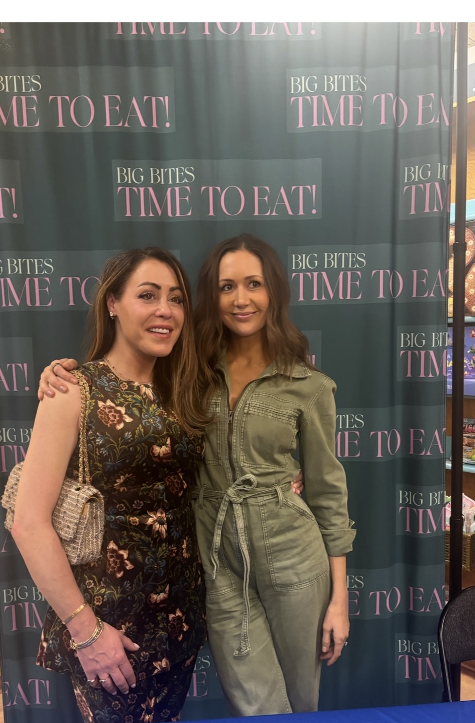 Two women pose and smile together in front of a green backdrop with the text BIG BITES TIME TO EAT repeated in pink and white letters. One woman wears a floral outfit; the other wears a green jumpsuit.