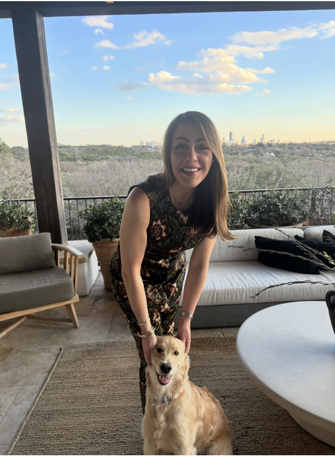 A smiling woman in a floral dress bends down to pet a happy golden dog on a patio with outdoor furniture, overlooking a scenic view of trees and a distant city skyline under a partly cloudy sky.