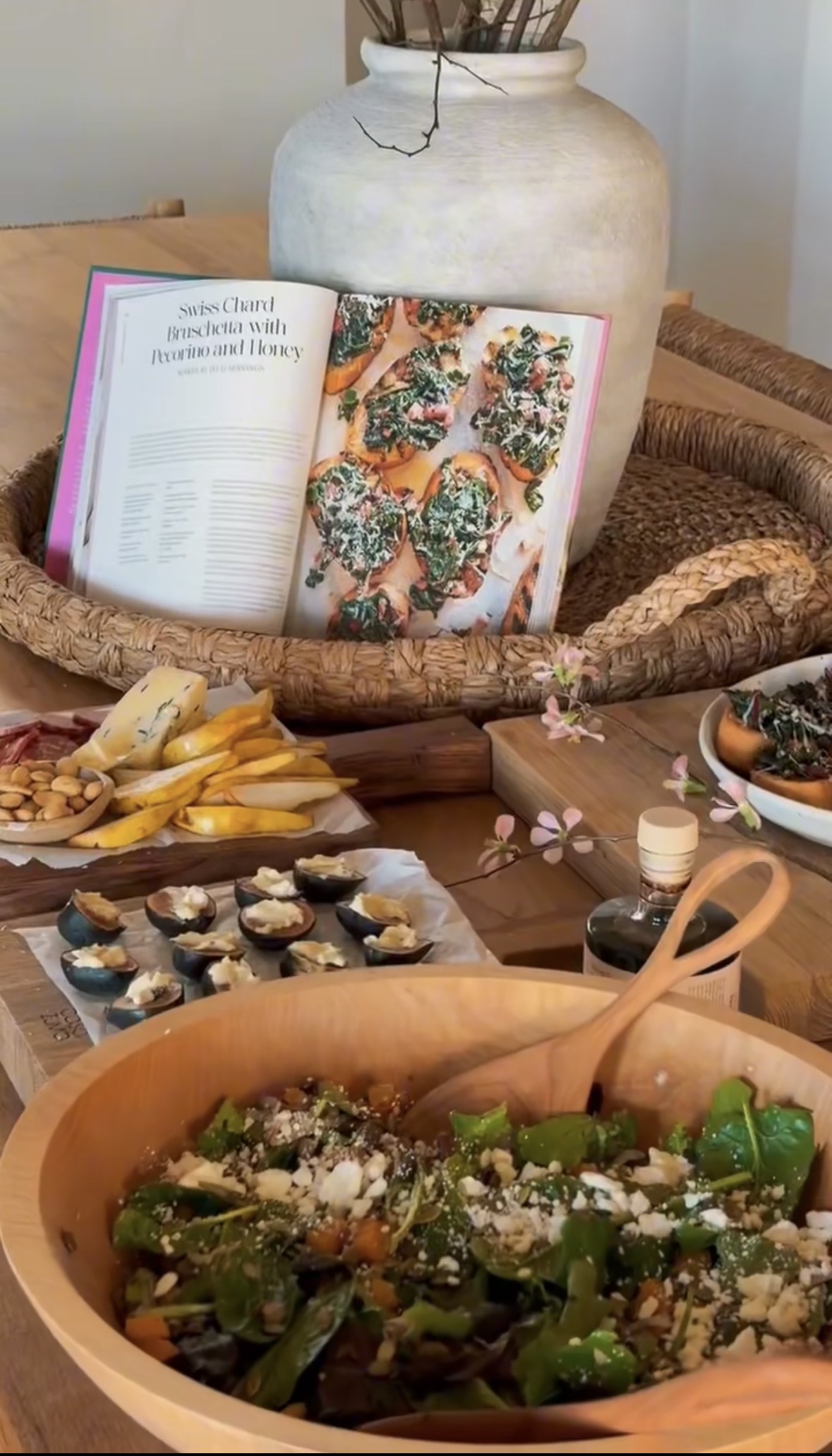 A table set with a wooden salad bowl, a cheese and fruit platter, small appetizers, and an open cookbook displaying a Swiss chard bruschetta recipe, all arranged near a large ceramic vase with branches.