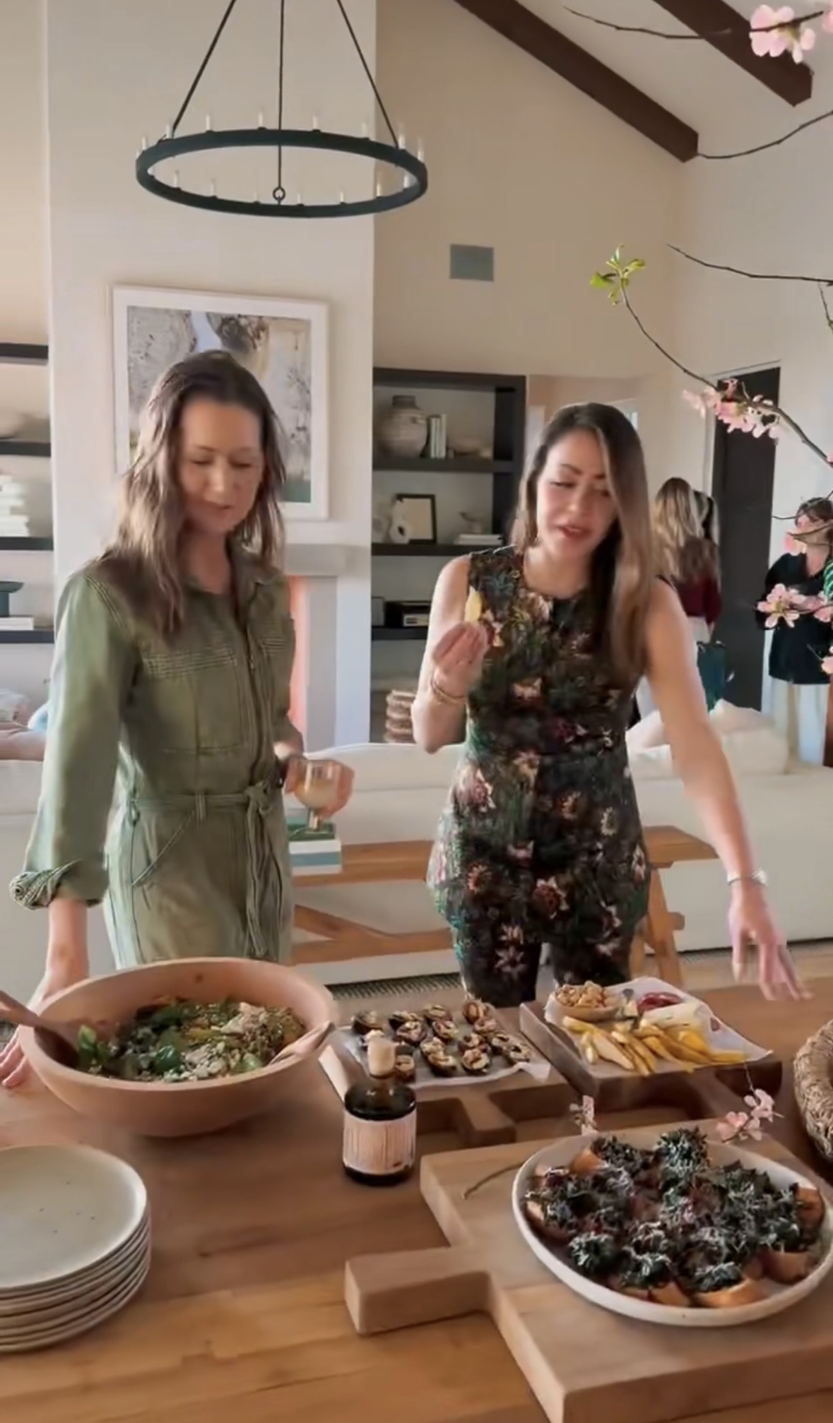 Two women stand by a dining table with various appetizers, salads, and snacks in a bright, modern living room. One woman eats, while the other looks at the table. Other people are in the background.