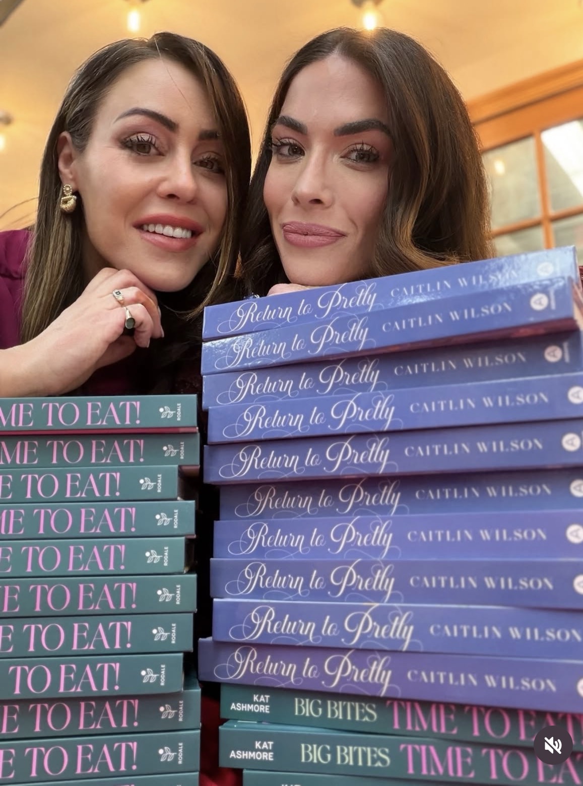 Two women smile and pose close together behind stacked hardcover books titled Return to Pretty and Time to Eat! with colorful covers on display.