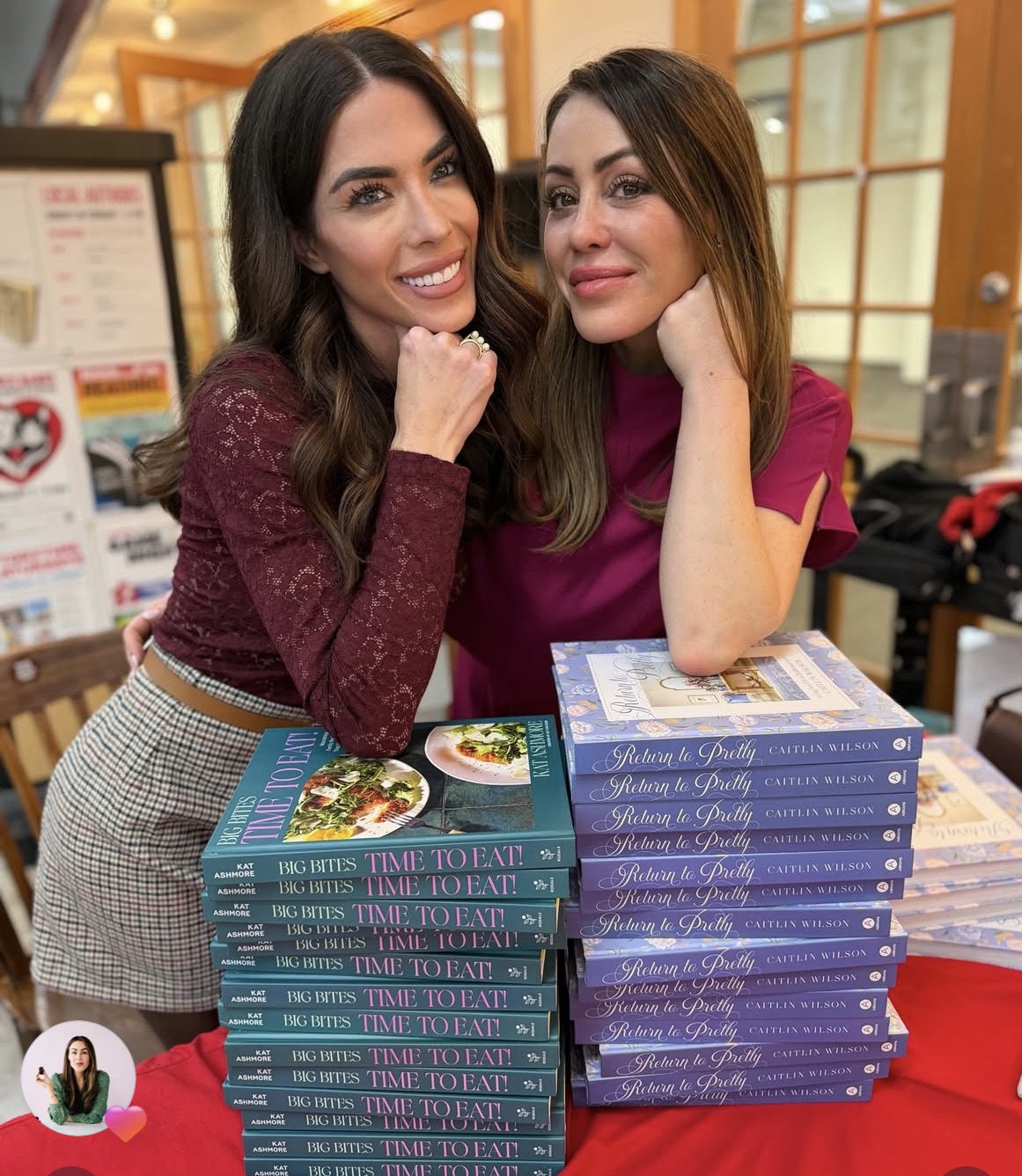 Two women smile at a table with stacks of books in front of them. The books include &ldquo;Big Bites: Time to Eat&rdquo; and &ldquo;Return to Pretty.&rdquo; They are indoors, posing with their heads resting on their hands.
