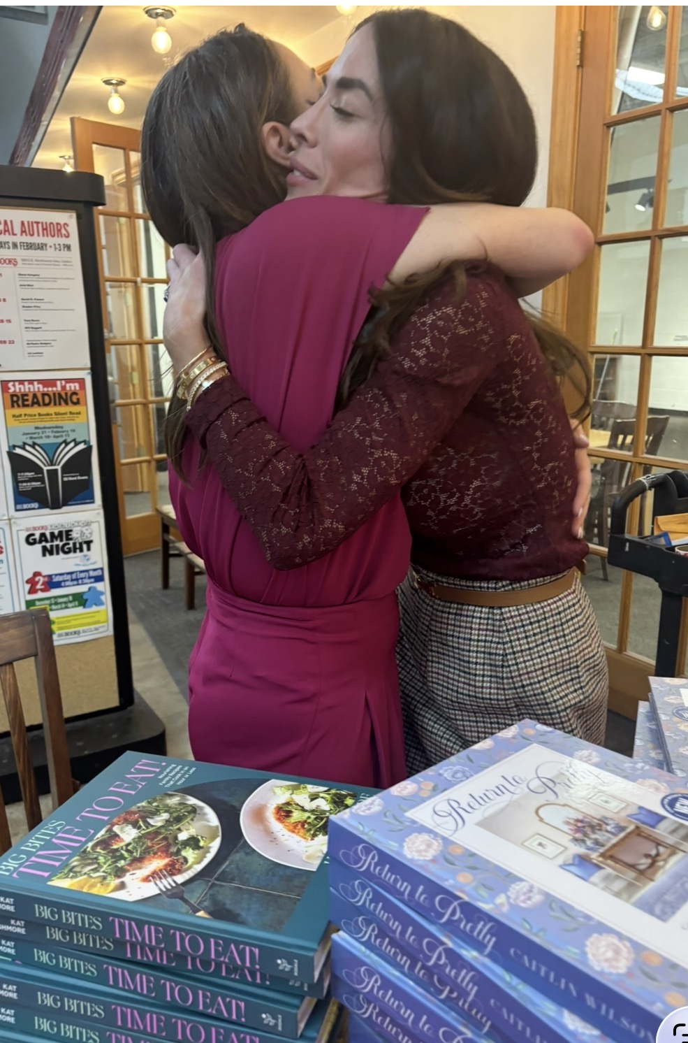 Two women embrace warmly at a bookstore event. Stacks of cookbooks are displayed on the table in front of them. A signboard with event information and book covers is visible in the background.
