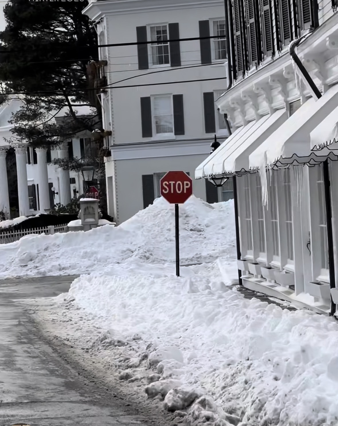 A stop sign stands on a snow-covered street with large snowbanks and buildings in the background. Icicles hang from an awning above white-framed windows, and another stop sign is visible further down the road.