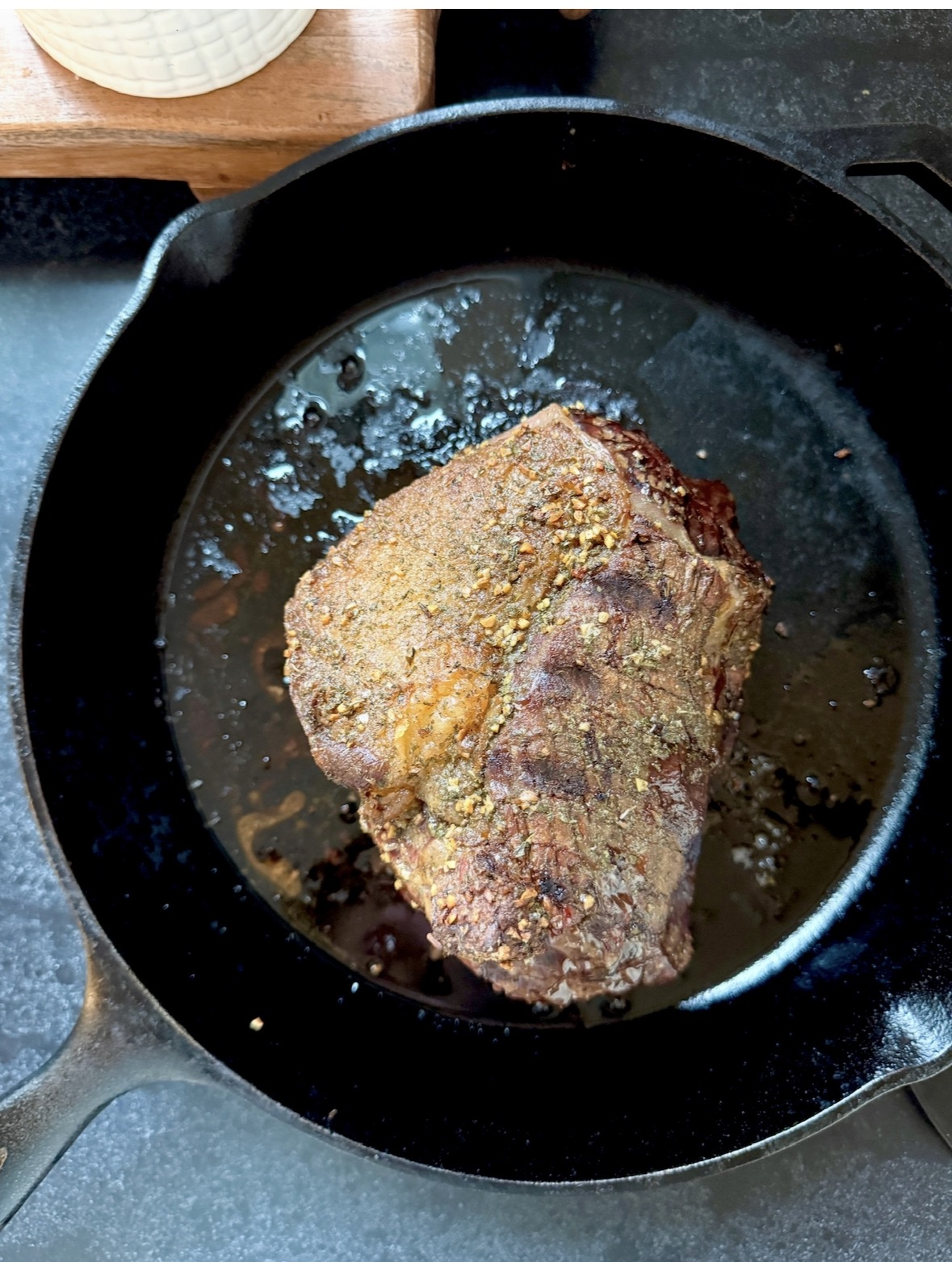 A seasoned piece of beef is searing in a black cast iron skillet, with browned edges and visible spices on the surface. The skillet sits on a dark countertop.