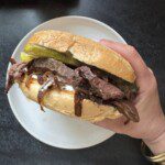 A hand holding a sandwich filled with sliced beef, pickles, caramelized onions, and sauce, with a crusty bun, over a white plate on a dark surface.