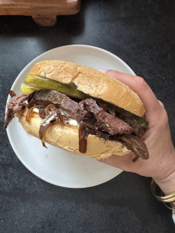 A hand holding a sandwich filled with sliced beef, pickles, caramelized onions, and sauce, with a crusty bun, over a white plate on a dark surface.