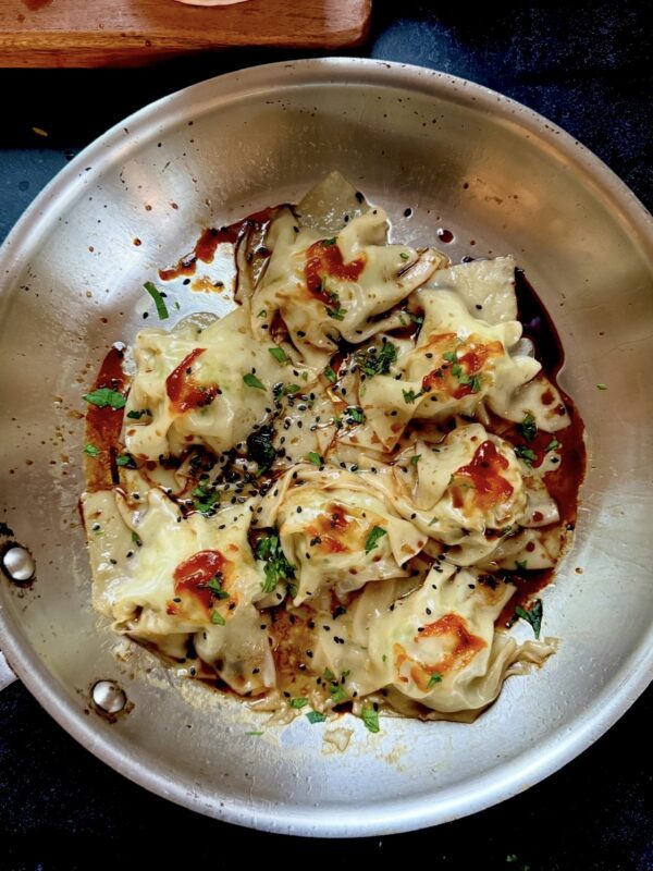 A stainless steel pan filled with dumplings garnished with chopped herbs and black sesame seeds, topped with a drizzle of sauce. The dumplings are lightly browned and arranged closely together.