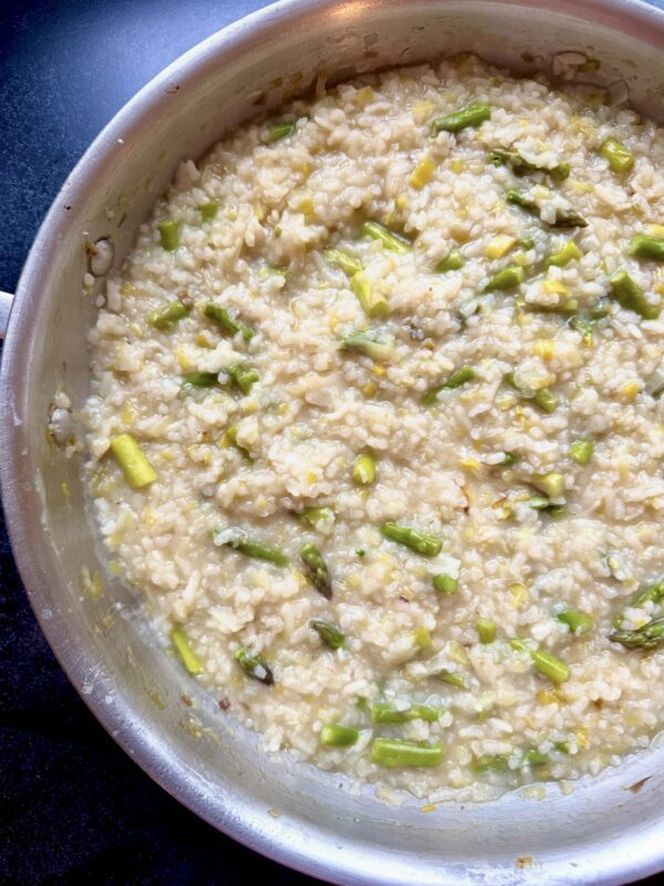 A close-up of a pan filled with creamy risotto, featuring pieces of green asparagus mixed throughout the rice. The dish has a slightly glossy texture, indicating it was freshly cooked.