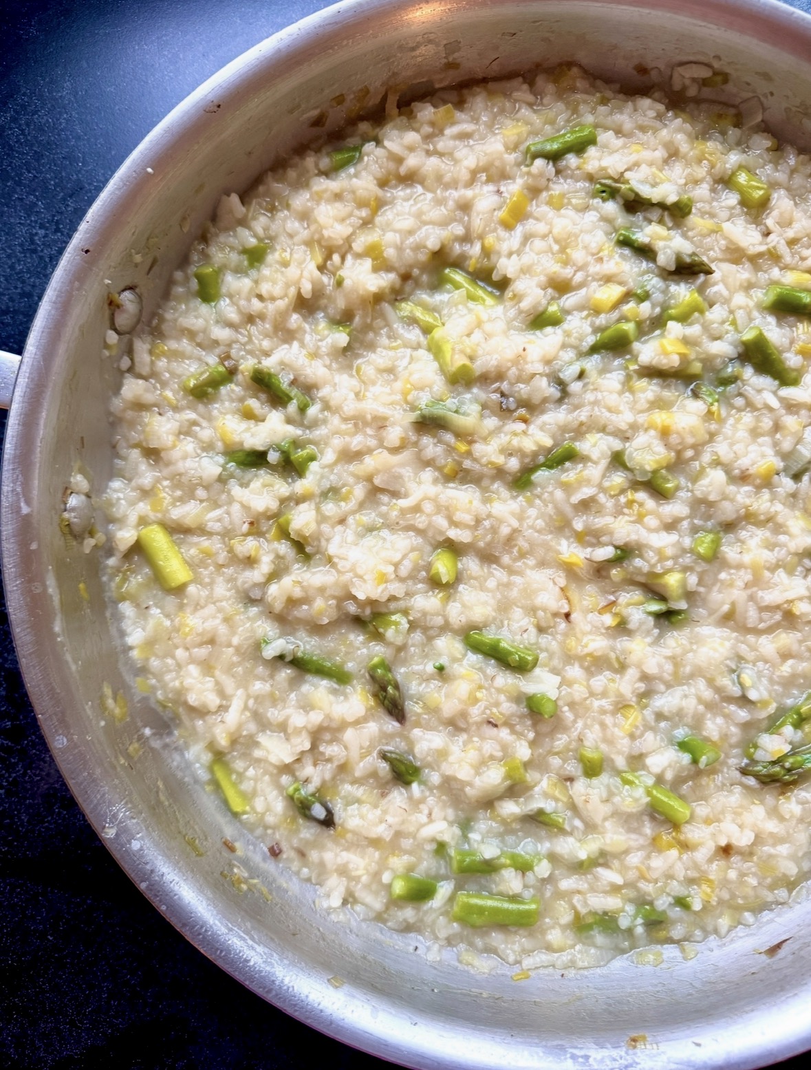 A close-up of a pan filled with creamy risotto, featuring pieces of green asparagus mixed throughout the rice. The dish has a slightly glossy texture, indicating it was freshly cooked.