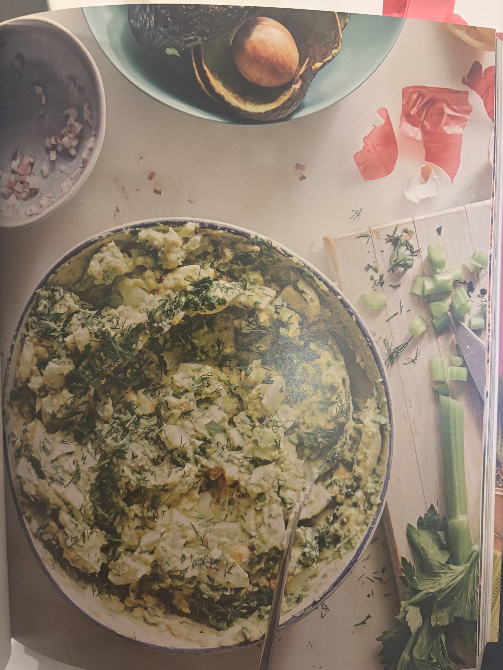 A bowl of creamy egg salad with chopped herbs sits on a table, surrounded by celery stalks, sliced avocado with pit, prosciutto, and a small dish of diced red onions. A spoon rests in the salad.