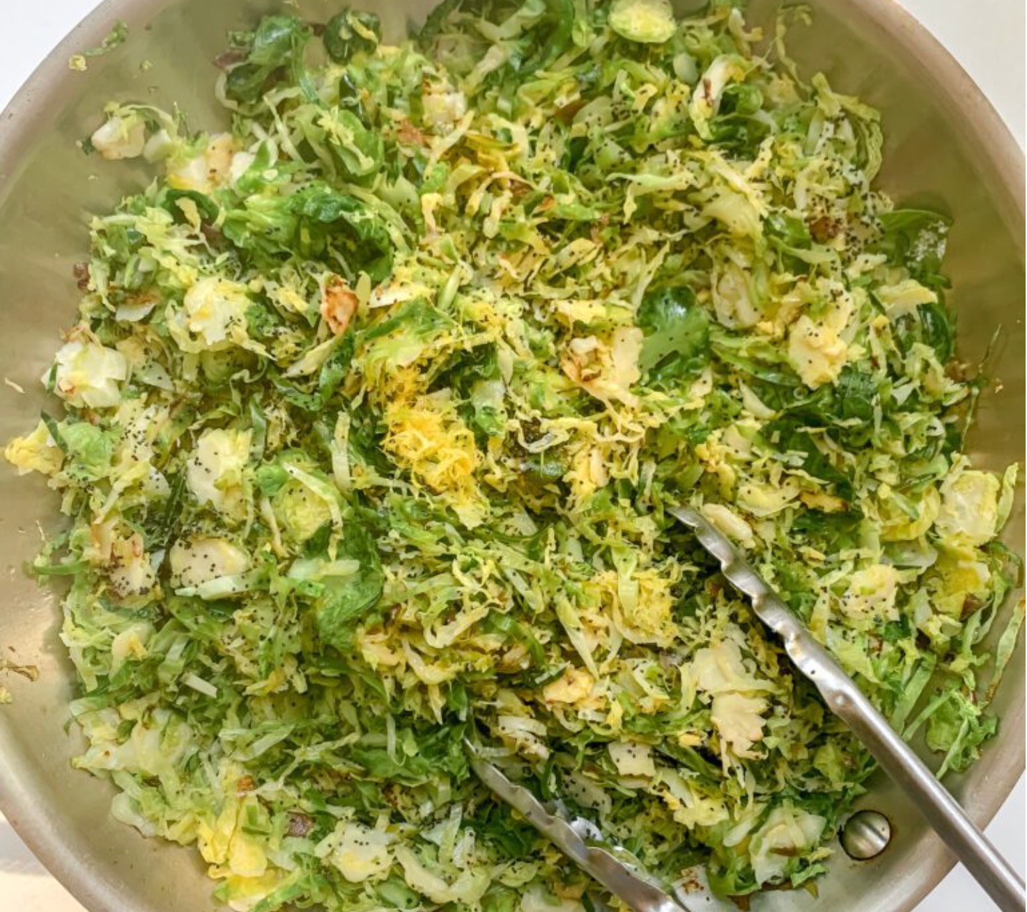 A pan filled with saut&eacute;ed, finely shredded Brussels sprouts, garnished with seasoning and lemon zest. Metal tongs rest on the edge of the pan.