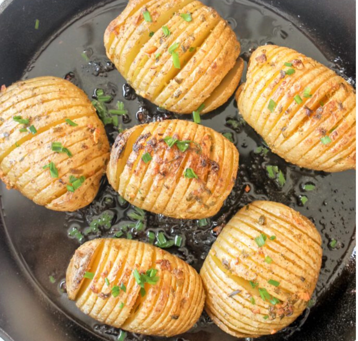 Six Hasselback potatoes with thin, evenly spaced slices are baked to a golden brown in a black skillet and garnished with freshly chopped chives.