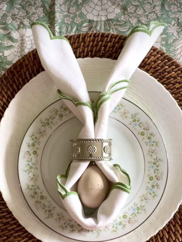 A place setting with a white napkin folded like bunny ears, held by a silver napkin ring over an egg, atop stacked floral plates and a woven charger, on a green patterned tablecloth sets the scene for My Easter Menu.