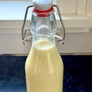 A glass bottle with a flip-top cap contains a creamy, pale yellow liquid. The bottle is set on a dark counter in front of a window with white trim.