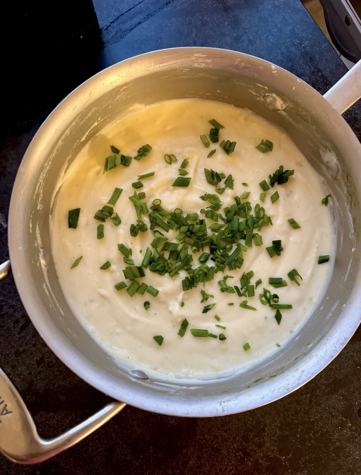 A pot of creamy mashed potatoes topped with freshly chopped chives sits on a dark countertop.