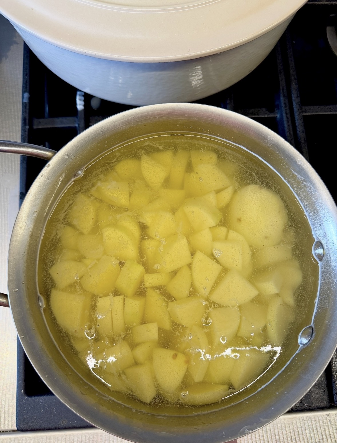 A pot of diced yellow potatoes boiling in water on a stovetop, with a white pot lid nearby in the background.