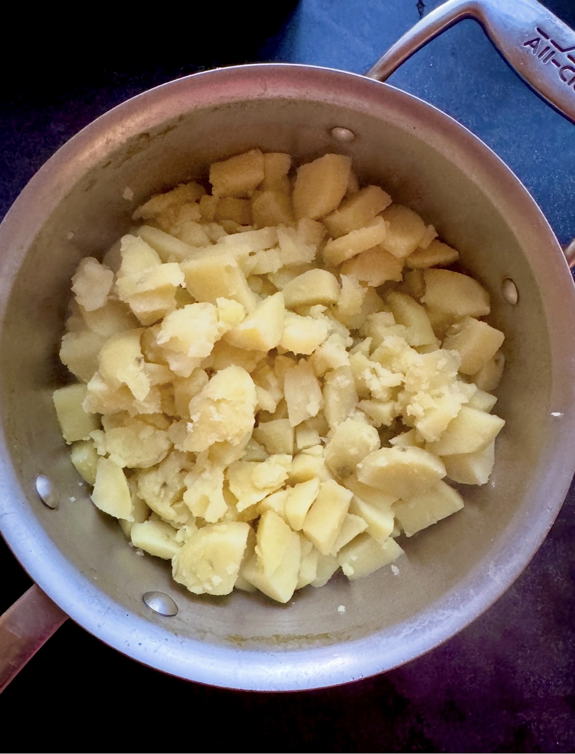 A metal pot filled with cooked, chopped, and slightly mashed potatoes sits on a dark countertop.
