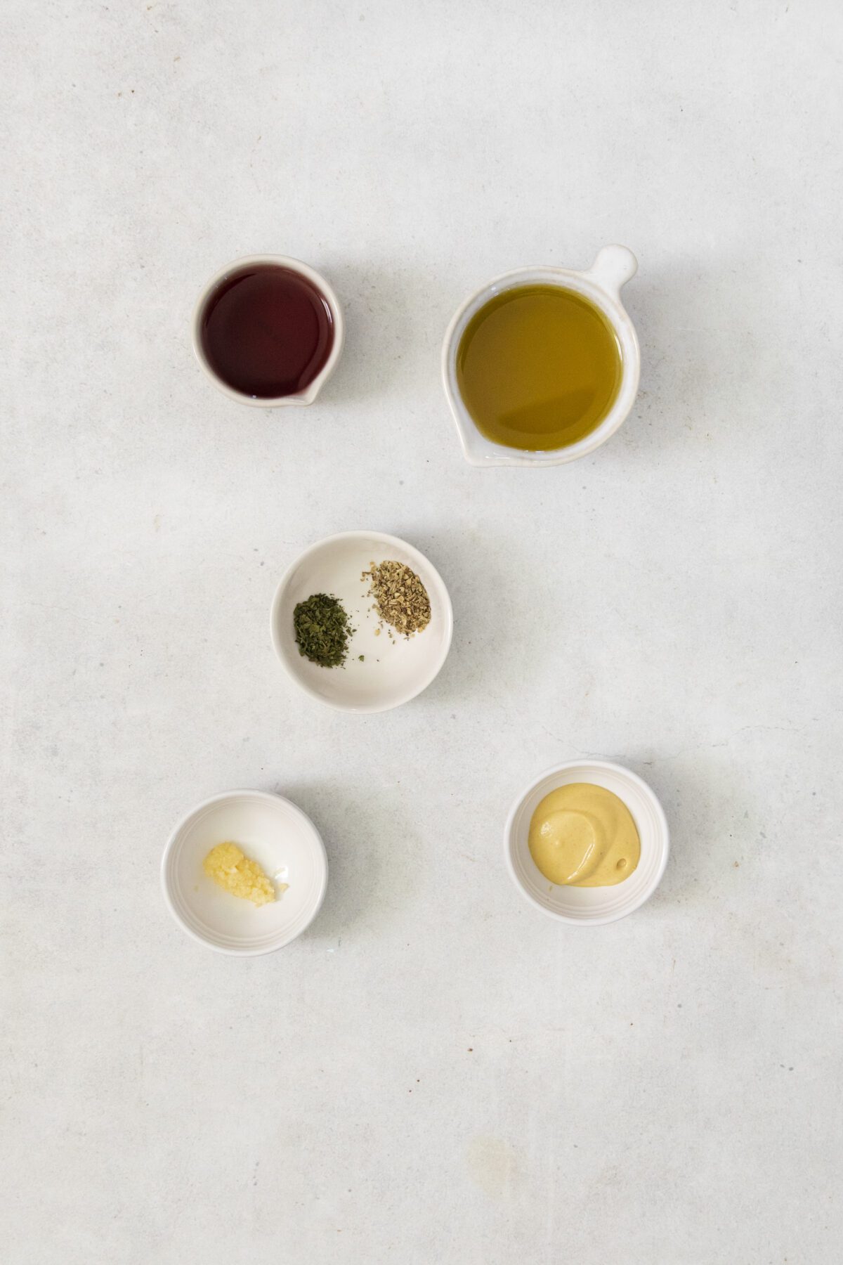 Five small bowls with ingredients on a light surface: one with red vinegar, one with olive oil, one with dried herbs, one with minced garlic, and one with Dijon mustard.