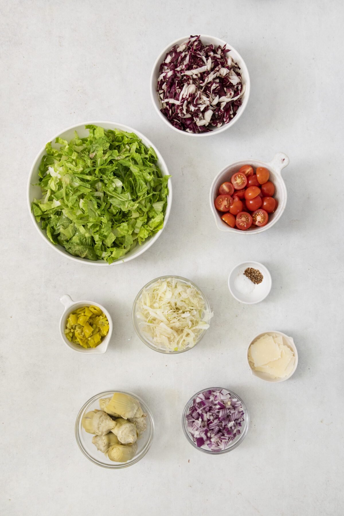 Bowls and cups filled with various salad ingredients, including chopped lettuce, shredded radicchio, grape tomatoes, diced pickles, shredded cabbage, chopped red onion, artichoke hearts, sliced cheese, and a pinch of seasoning.