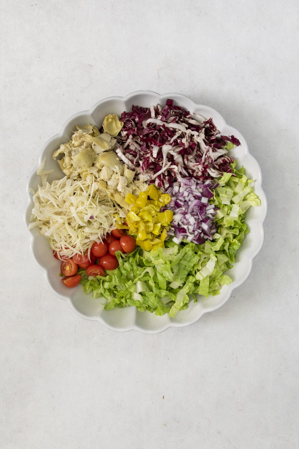 A scalloped white bowl filled with neatly arranged groups of chopped lettuce, cherry tomatoes, shredded cheese, artichoke hearts, red onion, radicchio, and yellow pepperoncini on a light background.