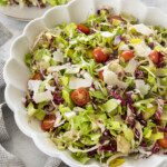 A large white bowl filled with a colorful chopped salad of lettuce, radicchio, cherry tomatoes, artichoke hearts, and shaved parmesan. A plate of salad and a bowl of cherry tomatoes are in the background.
