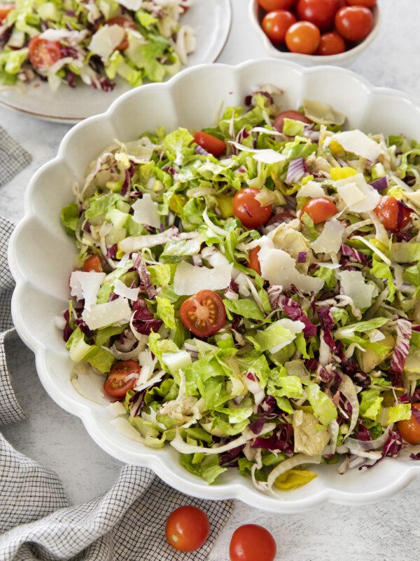 A large white bowl filled with a colorful chopped salad of lettuce, radicchio, cherry tomatoes, artichoke hearts, and shaved parmesan. A plate of salad and a bowl of cherry tomatoes are in the background.