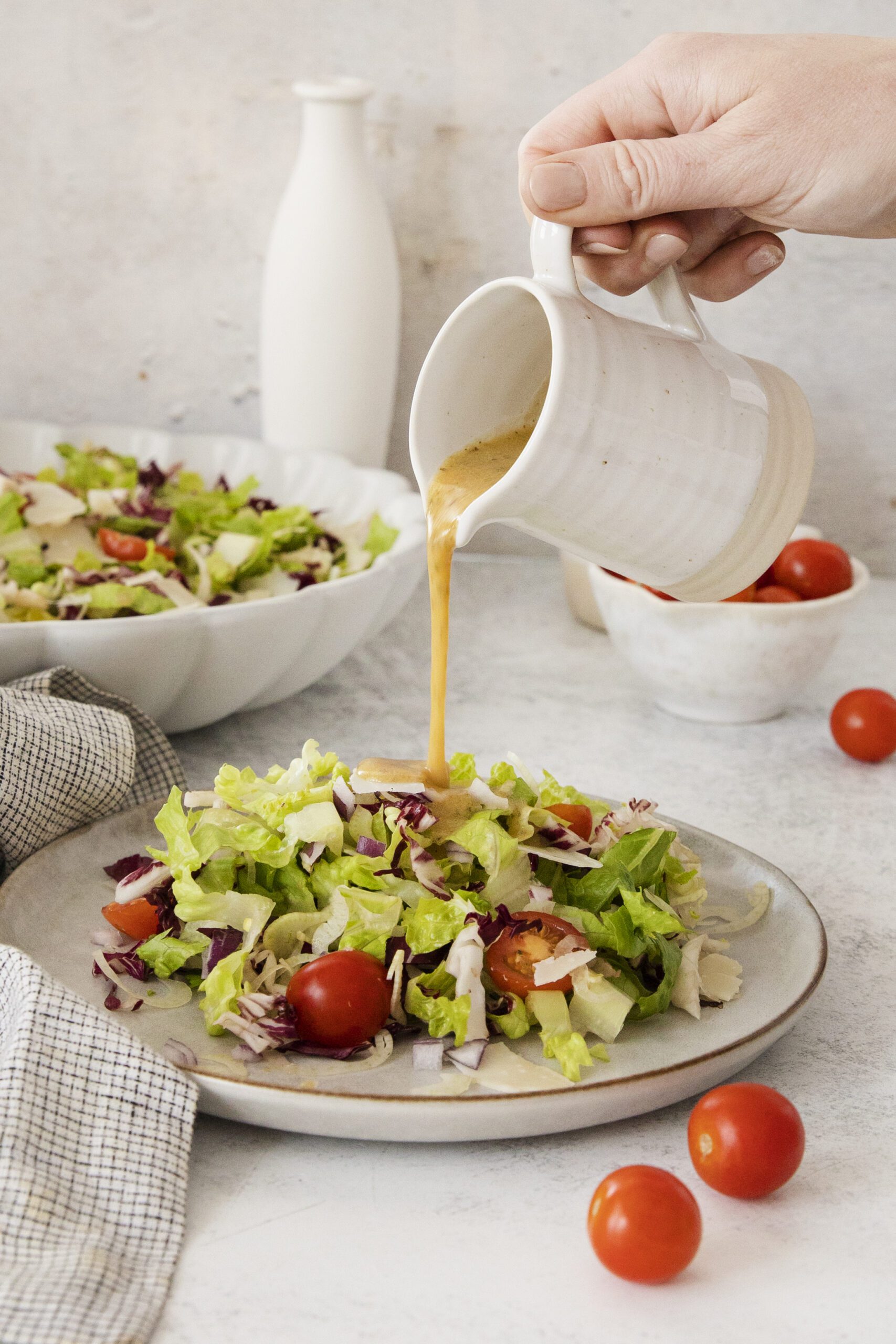 A hand pours dressing from a white pitcher onto a plate of fresh salad with lettuce, cherry tomatoes, and shredded vegetables. A bowl of salad, cherry tomatoes, and a white vase are in the background.
