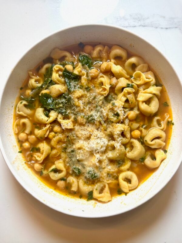 A bowl of tortellini soup with chickpeas, greens, and grated cheese.