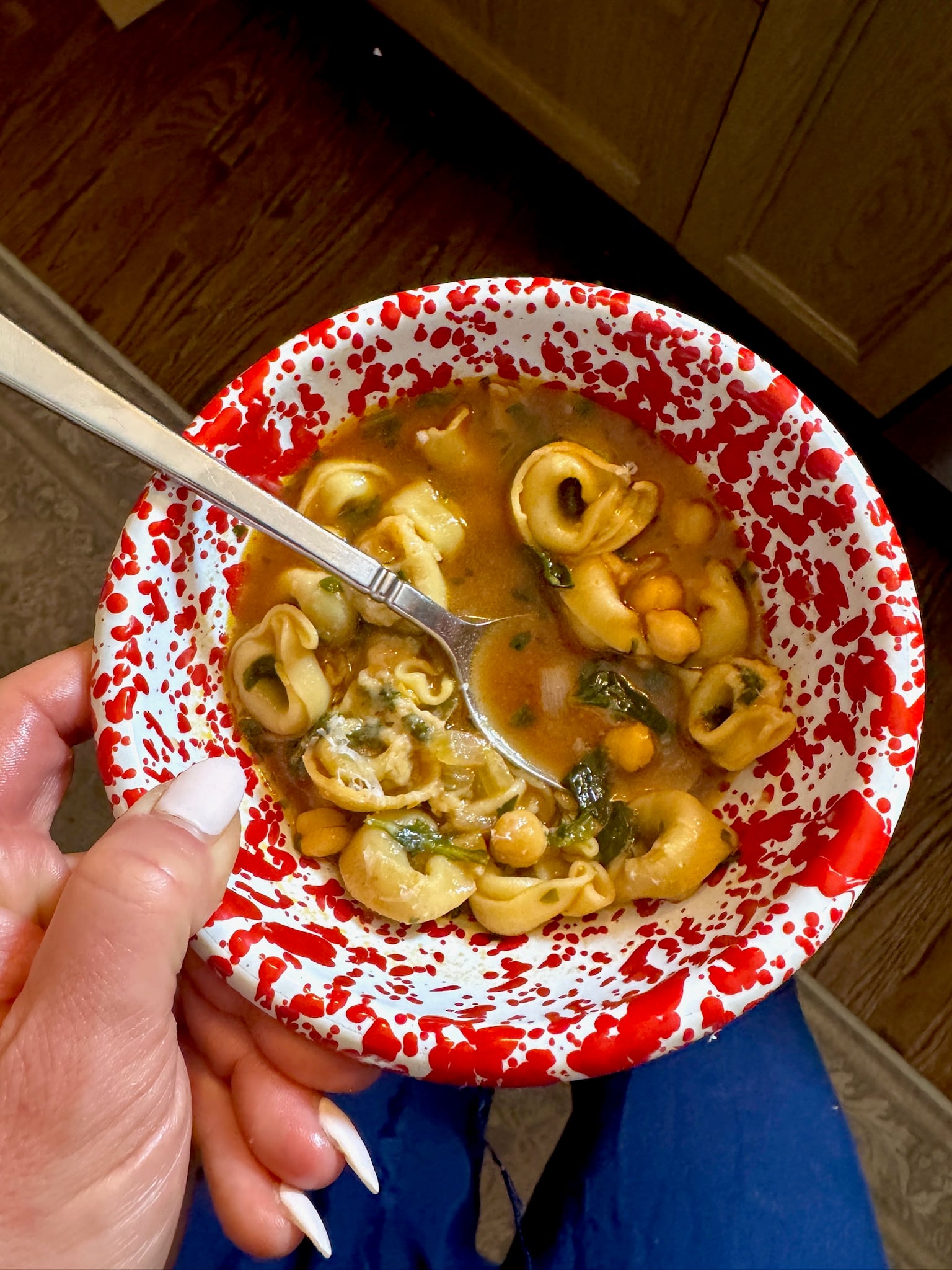 A hand holds a bowl of tortellini soup with chickpeas and greens in a red and white speckled bowl, with a spoon resting inside.