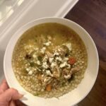 A bowl of chicken and pearl couscous soup topped with crumbled feta cheese and dill.