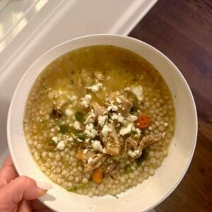A bowl of chicken and pearl couscous soup topped with crumbled feta cheese and dill.
