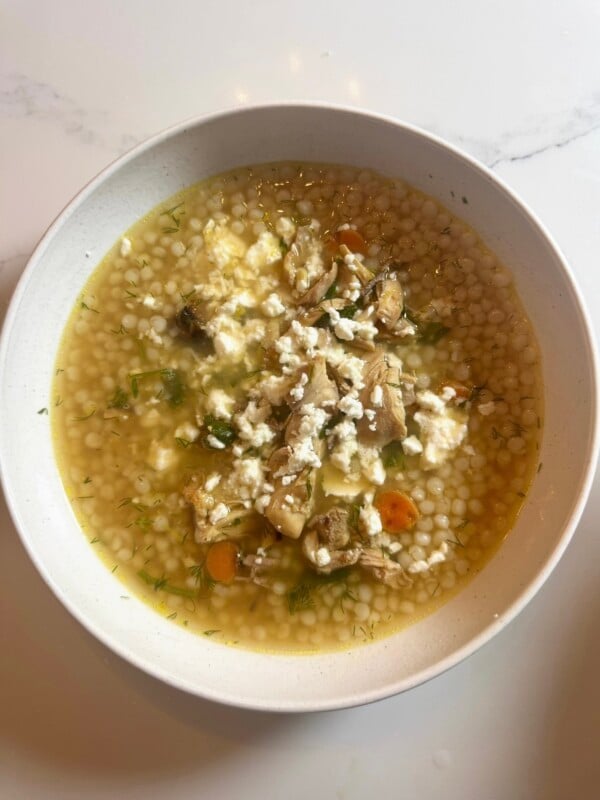A bowl of lemon chicken soup with Israeli couscous, topped with crumbled feta cheese and fresh dill.